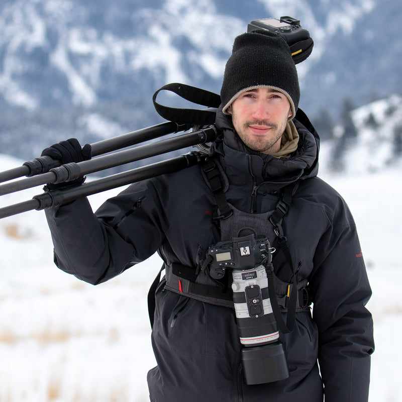 Connor Stefanison, professional nature photographer, standing in snow carrying a tripod and wearing a camera and lens in a Cotton camera harness with mountains in the background