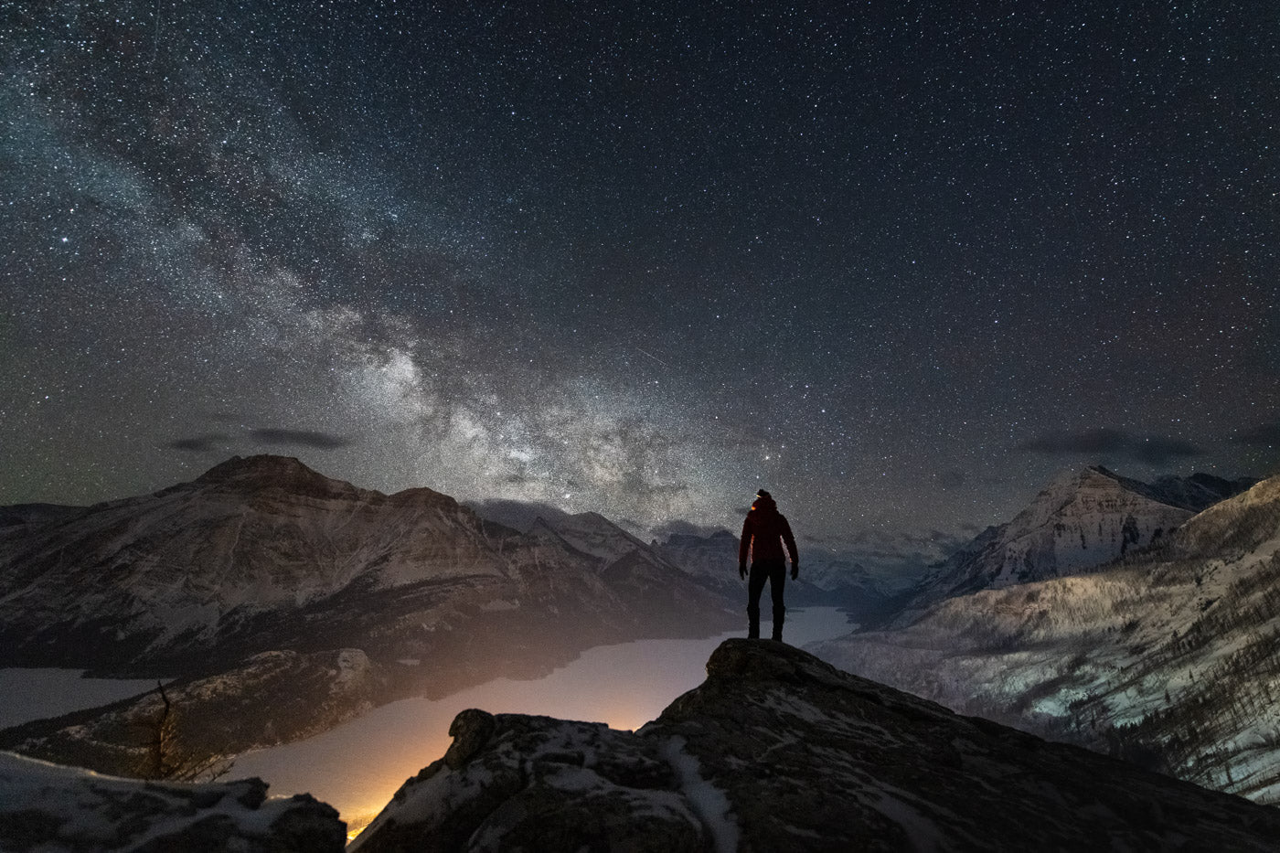 Professional photo taken of a person standing on a mountain peak at night looking at the dark sky, clouds, and stars